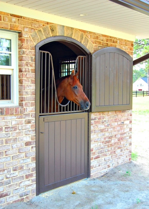 Gray vertical plank Dutch door with bay horse extending head over top section on red brick wall