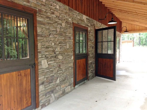 Covered barn breezeway with stacked stone walls and mahogany Dutch doors featuring metal grills