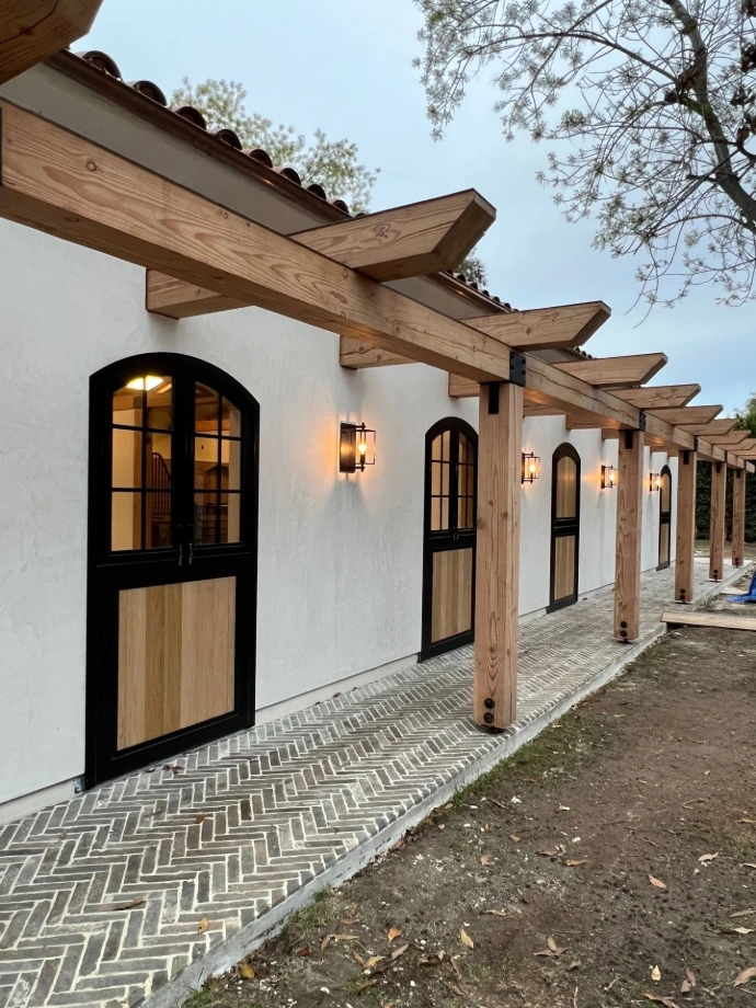 Evening view of covered barn aisle with illuminated arched black metal Dutch doors along white stucco wall