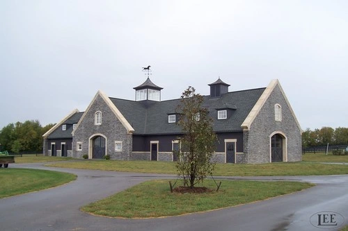 Gray stone barn complex with steep roofs and cupolas viewed from curved driveway