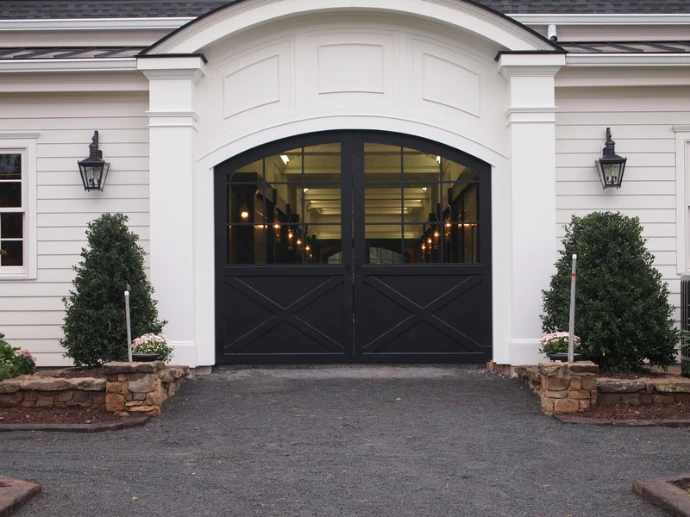 Black arched double doors on white barn with decorative shrubs and illuminated interior