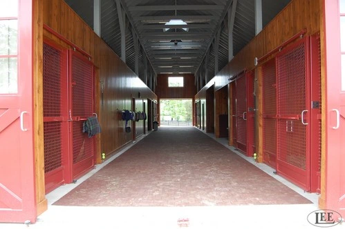 Long barn corridor with red metal stall doors featuring wire mesh panels on both sides
