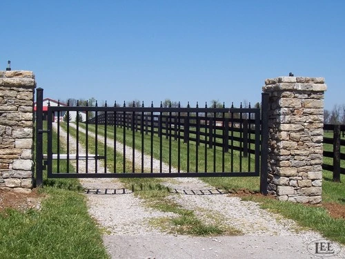 Black vertical picket gate spanning gravel path between stacked stone pillars under blue sky