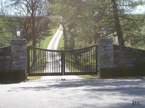 Black metal double gates partially open between stacked stone pillars on paved tree-lined driveway