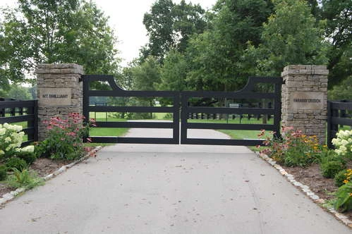 Black farm-style double gates with horizontal rails between stone pillars on paved entrance