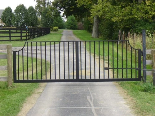 Black vertical bar swing gate across gravel driveway flanked by green pasture and trees