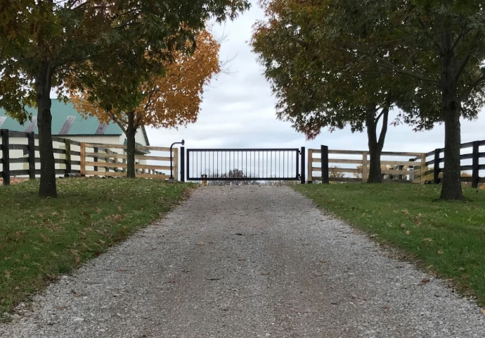 Simple black metal farm gate centered between wooden fence posts with autumn trees flanking gravel drive