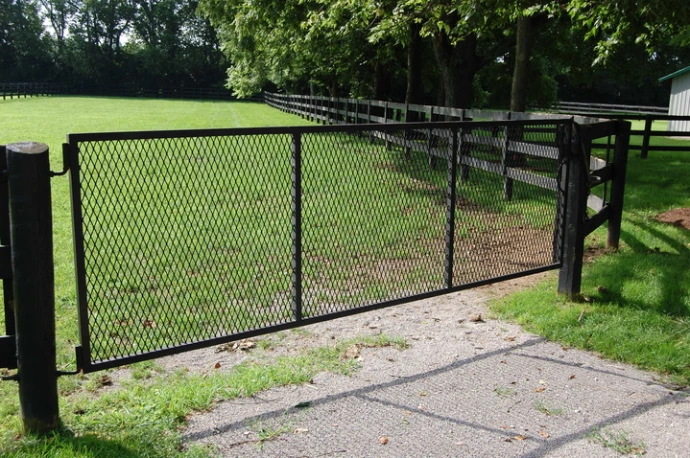 Black metal swing gate with chain link mesh panel partially open on gravel driveway leading to green pasture