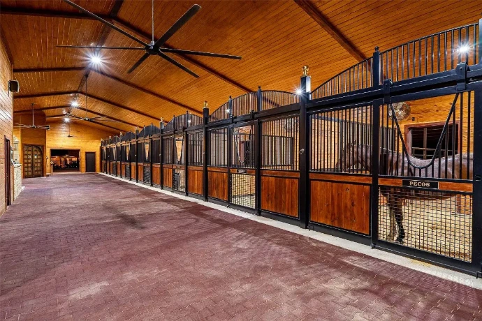 Long barn aisle with rich mahogany stall fronts, black vertical bar upper grills, burgundy flooring and horses visible in stalls