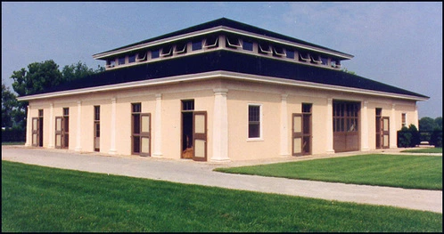 Cream colored barn with dark roof, arched stall openings and transom windows across front facade
