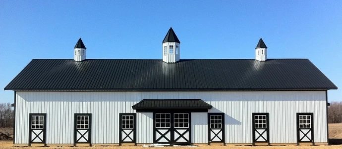 White vertical metal barn with dark roof, three cupolas and X-pattern stall doors