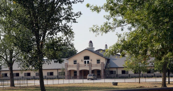 Tan stucco barn with stone accents viewed through trees on paved driveway