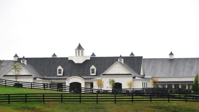 White barn complex with dark roofs, multiple cupolas and white board fencing